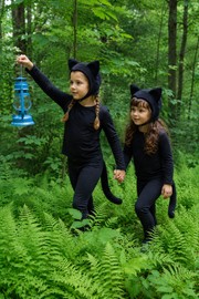 Two children in black cat costumes walking through a forest.