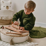 Child in a green sweater playing with wooden toys on a woven ottoman.