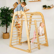 A large wooden climbing play structure with ladder bars, rope climbing, and a net panel, shown in a bright room with two children playing on it.