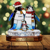 Stained glass penguin family ornament with 'Dave', 'Brenda', and 'Lucas' on a wooden base, Christmas tree in the background.