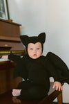 Baby in a black bat costume sitting on a piano bench.