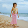 Young girl in a yellow top and pink skirt standing on a sandy beach with ocean and sky in the background.