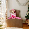 Child in a pink elf costume sitting on a chair with Christmas decorations in the background.
