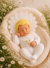 Baby in a white outfit and yellow hat with flower design, surrounded by flowers.