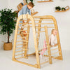 A large wooden climbing play structure with ladder bars, rope climbing, and a net panel, shown in a bright room with two children playing on it.
